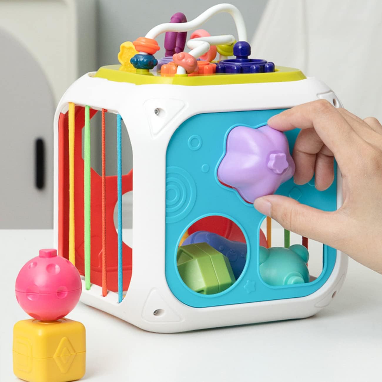Colorful baby cube toy with various shapes and a hand interacting with it on a white background
